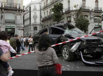 Unos viandantes contemplan el coche siniestrado colocado en la Puerta del Sol.