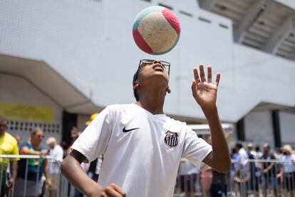 Un niño con la camiseta del Santos Fútbol Club juega con un balón, hoy, en las inmediaciones del estadio Vila Belmiro, en la ciudad de Santos (Brasil). El velatorio de Pelé, fallecido el pasado jueves a los 82 años, comenzó este lunes para que los aficionados brasileños rindan homenaje al mítico delantero, considerado por muchos el mejor futbolista de la historia. 