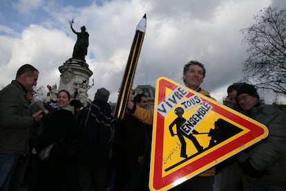 Um homem segura um lápis e uma placa com os dizeres "Vivemos todos juntos".