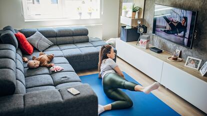 Uma mãe faz exercício diante da televisão enquanto seu bebê descansa no sofá.