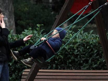 Una mujer fuma un cigarrillo ante un columpio ocupado por un niño en el parque del Retiro de Madrid