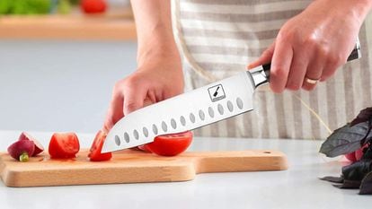 A person cuts some tomatoes using a Santoku-type knife, on a wooden board.