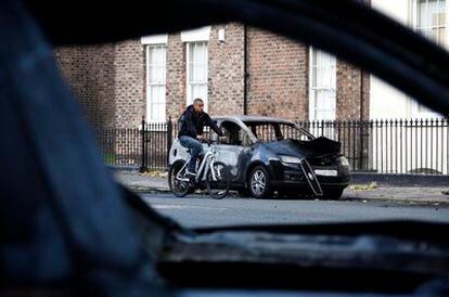 Un joven pasea en bicicleta junto a los restos de un coche incendiado